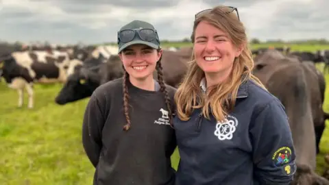 Emily and Georgie Paul standing in a field with cows. They are wearing dark jumpers and both are smiling.