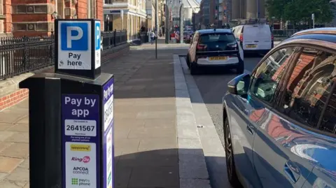 A parking metre on a street showing a P sign with text saying pay here. Two cars are parked in the on-street bays.