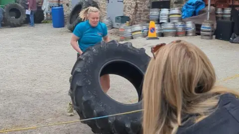 Chelsea Shoesmith A woman in a blue t-shirt and shorts is flipping a large tractor tyre. She is doing so in a pub car park filled with barrels and other tractor tyres.