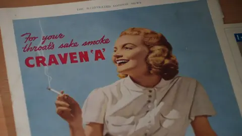 Shaun Whitmore/BBC An advertisement for Craven A cigarettes showing a colour drawing of a blonde woman wearing a white shirt and with a cigarette in her right hand. It says for your throat's sale smoke Craven A. It comes from the Illustrated London News.