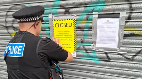A police officer attaches a yellow closure notice to a shutter outside a shop.