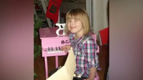 Cambridge University Hospitals Grace aged three sits in front of a pink toy piano. She wears a pink and blue checked dress.