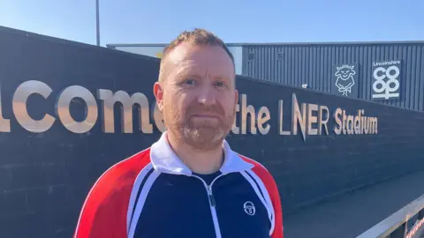 Sean Dunderdale/BBC A head and shoulders picture of Gary Hutchinson standing outside a football stadium, smiling at the camera. He has short ginger hair and facial hair. He is wearing a blue, red and white zip-up jacket. Behind him is a black wall that reads "Welcome to the LNER Stadium" in larger silver letters. The sky is blue.