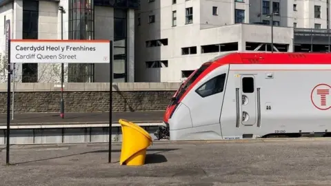 A train stopped at a railway station, next to a sign that says Cardiff Queen Street in English and Welsh, and a bin.