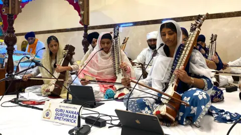 Three women on a stage playing Indian string instruments. In front them are small microphones on thin stands, and they are looking at ipads which sit on the floor. There is a sign in front of one of the women that says Guru Nanak Gurdwara Smethwick