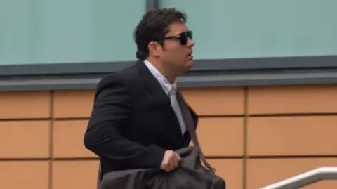 A man with black hair walking into court with a duffle bag around his shoulder. He's wearing a black suit and white shirt. The building behind him is orange/brown brick.