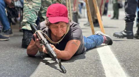 Nicole Kolster/BBC Kırmızı beyzbol şapkası giyen yaşlı bir kadın, yerde yatarken bir tüfek tutmak için mücadele ediyor. Tüfek asfalta işaret ederken yere bakıyor. Arka planda kamuflaj üniformaları ve askeri botlar giyen birkaç kişi var. 
