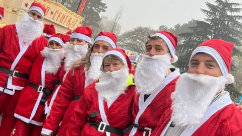 There are seven people dressed in Santa costumes wearing red suits, white fluffy beards and red Santa hats. 