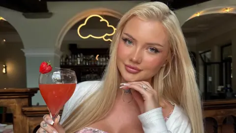 Handout A woman with long blonde hair is smiling and holding a cocktail. She is wearing a pink dress and white cardigan.