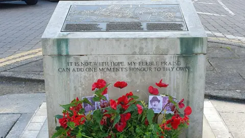 Soham Museum A light stone memorial with red poppy flowers around it and black and white pictures of four men arranged amongst the petals, behind it is a road way with double yellow lines.
