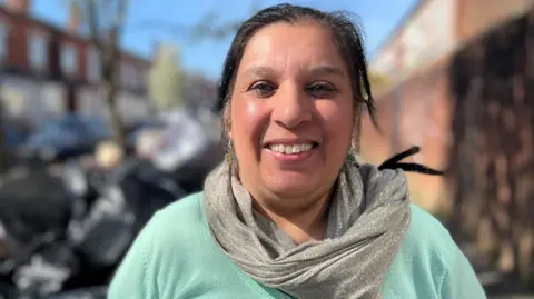 A woman stands on a street on a sunny day. She is wearing a pale green top and a silver scarf. She has straight black hair that is tied back.