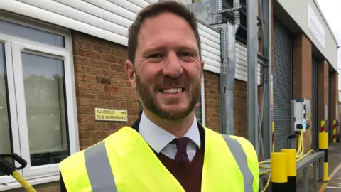 Matthew Gudgin/BBC James McCluskey, a man in a white shirt, a red tie and red jumper. He is wearing a fluorescent hi-vis jacket and is looking into the camera and smiling. 