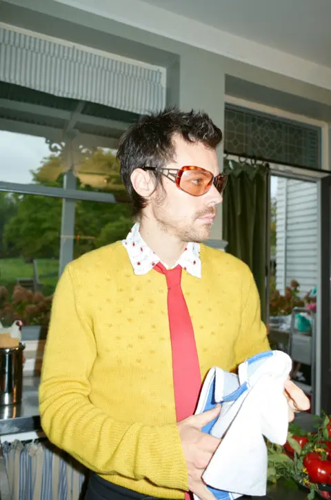 Sony Music Harry Styles in a yellow top, bright red tie and orange sunglasses, apparently holding a tea towel, in a promotional image