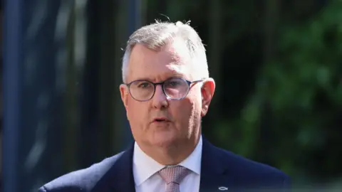 A man with short grey hair and rounded glasses is standing outside, with some out of focus trees behind him. The man is dressed in a dark suit jacket with white shirt and light pink tie, which has a textured pattern. There is a small silver fish symbol attached to the lapel of his jacket.