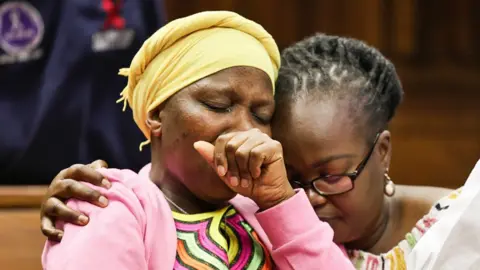 Luba Lesolle/Gallo Images/Getty Images A woman in glasses hugs Ntombizodwa Samane, the mother of Amantle Samane who wears a yellow headscarf and pink fleece over a yellow, pink, green and orange swirly striped top. Ms Samane holds her hand to her mouth and has her eyes closed in the Johannesburg High Court, South Africa - Monday 1 September 2025.