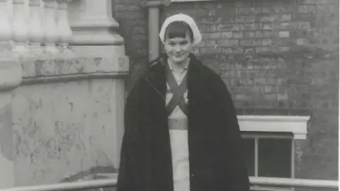 NHS Somerset Image shows black and white photo of Dawn Wintle smiling into the camera, with a brick-building in the background, and wearing her uniform. She wears a black coat with her hair pulled back and a white nurse cap.