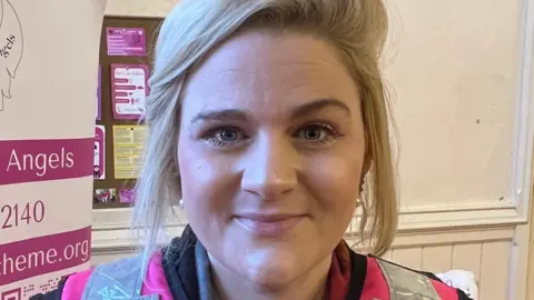 Minister for victims Alex Davies-Jones is wearing a pink Hi-vis jacket and has short blond hair. She is smiling at the camera.