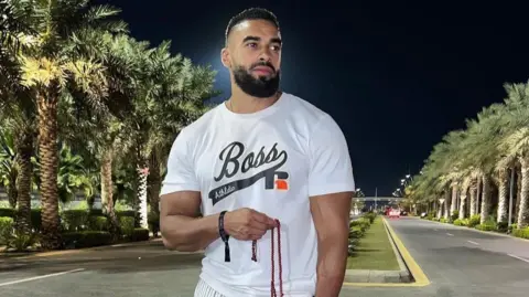 Handout Latif Ayodele in a white t-shirt standing in front of a road lined with palm trees.