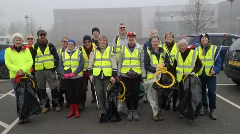 The image shows 15 people wearing hi-vis jackets and holding empty bin bags. Fog, a building and a few trees can be seen behind them.