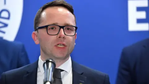 PA Media Dáire Hughes speaking at a microphone. He has dark hair and is wearing glasses. He has a navy suit and black tie.