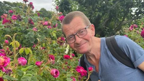 A picture of Stephen Allen, who is wearing a blue T-shirt and a black rucksack on his back, leaning towards pink roses.