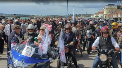 A huge group of motorcyclists are gathered by the sea in Bournemouth. Many of the bikers are smartly dressed and wearing ties. 