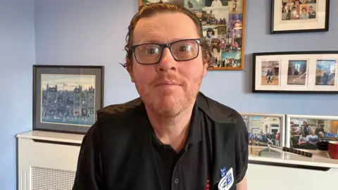 Adrian. He is wearing a black t-shirt and glasses with thick black frames. He has short dark hair. He is leaning forward slightly, looking straight at the camera. 