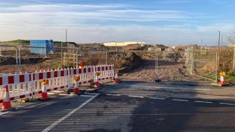 A closed road, complete with white and red barriers to the left, a fenced off area, with metal barriers, with a mud track, cones and telegraph poles in the distance. 
