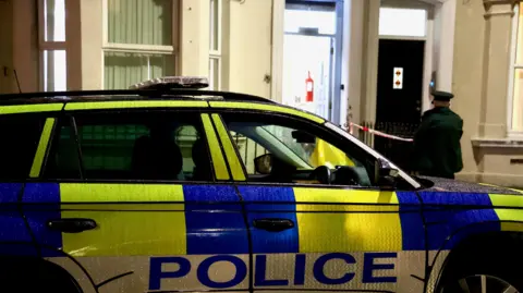 A police car outside a cream house. The door is open and an officer standing outside. The car is green white and blue. 