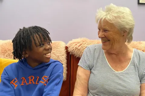 Smiling young black boy with dark hair sits beside smiling older white woman with grey hair