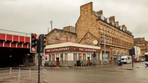 A Glasgow street, with a pub on the corner, and a large sandstone building to one side. Just behind it is a railway bridge.