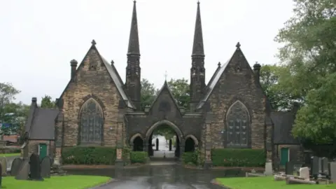 Geograph/David Morris Two gothic buildings with large arched windows are connected by an ornate stone archway. In the background are two large towers which come to a point