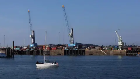 BBC St Peter Port Harbour. There is a small boat in the sea and in the background there are cranes and various shipping containers. 