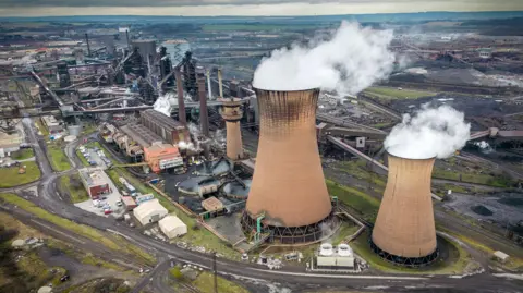 Christopher Furlong / Getty Images Steam comes out of huge towers in a wide shot of Scunthorpe steel works against a cloudy sky in 2023.