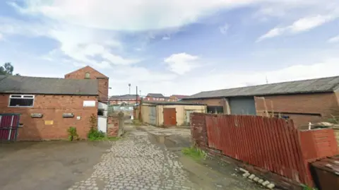 A small car park with cobblestones and tarmac and some red brick buildings around with fencing to one side 