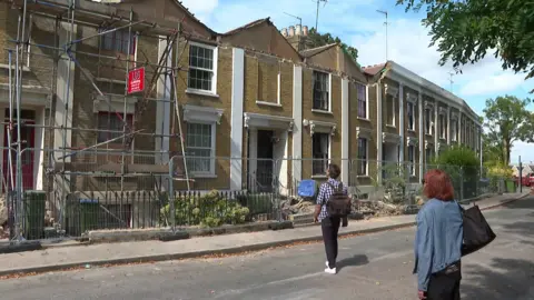 A row of terraced houses on Woodland Terrace in Charlton behind temporary fencing after part of the roofline and frontage collapsed. Two people stand outside the properties, where scaffolding has been erected and debris is visible along the pavement.