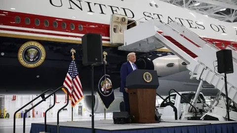 Trump standing at a podium on a stage in front of the new jet, painted white with dark red and blue stripes. There is a presidential seal and the words "United States of" visible. 