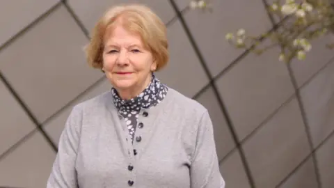 Standing in the Arc shopping centre, Joanna is wearing a grey cardigan and black and white patterned scarf. 
