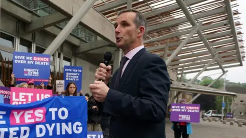 McArthur standing outside the Scottish Parliament speaking into a microphone. Behind him are protesters in favour of the bill, holding a banner and placards.