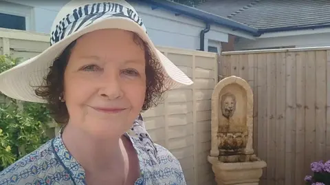 Paul Burnell/BBC Lesley Middleton has a broad brimmed sunhat, flowered top and red hair. IN the bsackground is a bush and fountain.