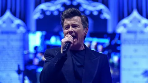 A man in a dark suit sings into a microphone on a stage. Behind him the area is lit in blue