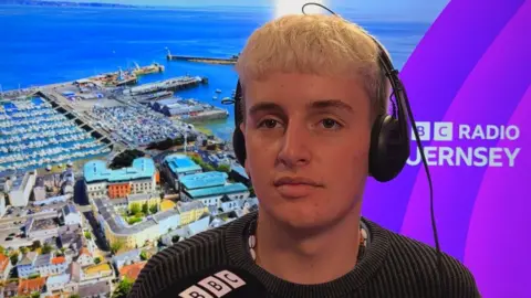Zack lleva un jersey oscuro y tiene el pelo rubio brillante. Está sentado en un estudio de radio de la BBC en Guernsey y lleva puestos unos auriculares.