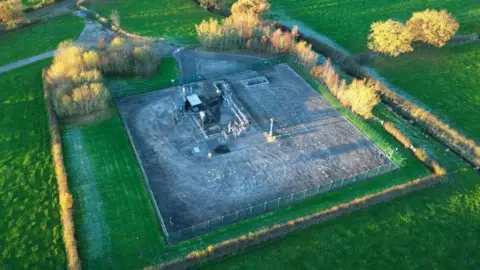 An aerial shot of a small square grey facility surrounded by trees and green fields.