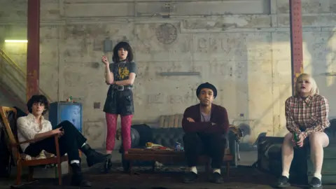 BBC/BANIJAY RIGHT/KUDOS A scene from This Town, with four characters standing in a warehouse which has a sofa and several chairs. Two women and two men are in the scene.