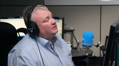 Stephen Nolan in a BBC radio studio.  He has short, grey hair and is wearing headphones and an open-necked blue shirt.   He is speaking into a blue microphone and there are several computers around him in the studio. 