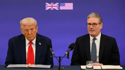 Trump and Starmer sit at black desk with black microphones. A purple coloured screen behind them has small UK and US flags pressed into it. Starmer is wearing a black suit and patterned tie with a white shirt. Starmer is looking ahead with his hands crossed over one another. Trump is looking straight ahead and appears to be scowling. He is wearing a navy suit with a red tie and a white shirt.