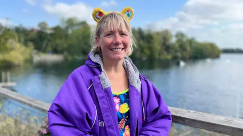 A woman wearing a purple waterproof coat over a multicoloured swimming costume. She's wearing a pair of yellow Pudsey Bear ears and smiling at the camera with a lake in the background.