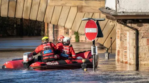 PA Media A small boat with three people in red hi-vis jackets and helmets, in a flooded area with half a road sign sticking up out of the water.