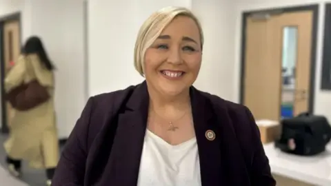 Fiona Kane is smiling at the camera. She has short blonde hair. She is wearing a plum coloured blazer with a white t-shirt underneath. She is standing in a school corridor.
