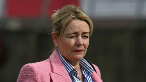 Getty Images A woman with blonde hair pulled back looks off to the side with a serious expression, wearing a pink tweed blazer over a blue and white striped shirt. A lanyard around her neck reads 'jobs, pay, conditions' in visible text. The background is blurred